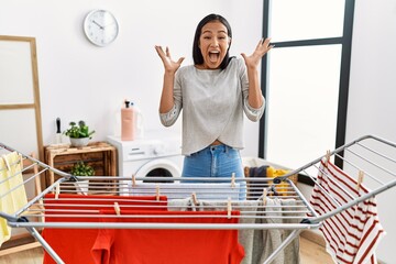 Wall Mural - Young hispanic woman putting fresh laundry on clothesline celebrating crazy and amazed for success with arms raised and open eyes screaming excited. winner concept