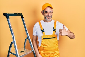 Poster - Handsome middle age man with grey hair holding ladder doing happy thumbs up gesture with hand. approving expression looking at the camera showing success.