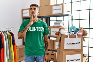 Sticker - Young handsome hispanic man wearing volunteer t shirt at donations stand thinking concentrated about doubt with finger on chin and looking up wondering