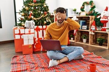 Sticker - Arab young man using laptop sitting by christmas tree covering eyes with hand, looking serious and sad. sightless, hiding and rejection concept