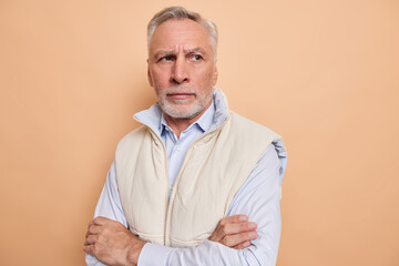 Wall Mural - Indoor shot of thoughtful elderly man keeps arms folded thinks of personal problems lost in thoughts remembers and misses something wears formal shirt and vest isolated over beige background