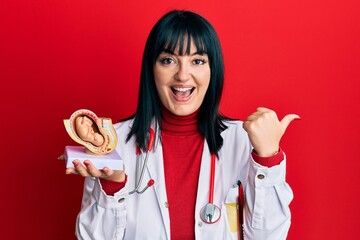 Sticker - Young hispanic gynecologist woman holding anatomical model of female uterus with fetus pointing thumb up to the side smiling happy with open mouth