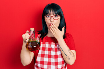 Wall Mural - Young hispanic woman wearing cook apron holding olive oil can covering mouth with hand, shocked and afraid for mistake. surprised expression