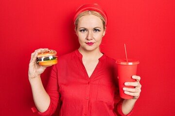 Canvas Print - Young blonde woman eating a tasty classic burger with fries and soda relaxed with serious expression on face. simple and natural looking at the camera.