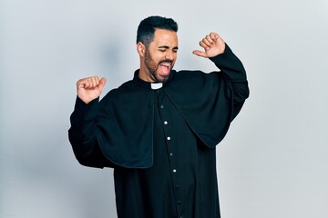 handsome hispanic man with beard wearing catholic priest robe dancing happy and cheerful, smiling mo