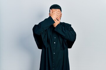 Poster - Handsome hispanic man with beard wearing catholic priest robe covering eyes and mouth with hands, surprised and shocked. hiding emotion