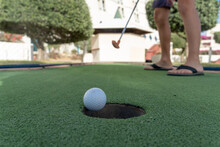 Young Child's Feet On A Minature Or Mini Golf Hole Close Up With Ball Falling To The Hole On Green Artificial Turf Outdoors.