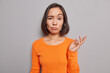 © wayhome.studio  - Studio shot of indignant Asian woman raises palm with hesitation cannot make decision dressed in casual orange jumper poses against grey studio wall looks in bewilderment hears something strange