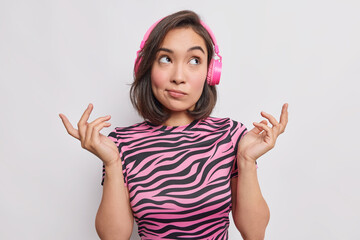 Wall Mural - Maybe yes or no. Hesitant young Asian woman shrugs shoulders has clueless expression makes choice daydreams while listening music wears casual striped t shirt isolated over white background.