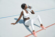 © gstockstudio - Beautiful young African woman in sports clothing drinking water while sitting on sports track outdoors
