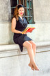 © Alexander Image - Sexy Woman Reading Outside. Wearing a black sleeveless trench coat dress, a hair band,  a young beautiful woman is sitting by a window, smiling, reading a red book..
