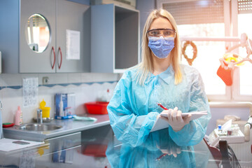 Portrait of female dentist in a protective suit, shield, mask and eyeglasses writing notes to planner book at dental office.