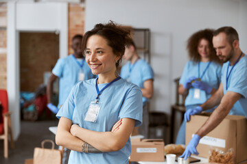 Wall Mural - Portrait of happy young woman, volunteer in blue uniform smiling away while standing with arms crossed indoors. Team sorting, packing items in the background