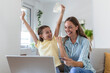 © Graphicroyalty - Low angle of happy small daughter with arms raised rejoicing while smiling mother in casual clothes focusing on screen and using laptop for online shopping in light modern apartment