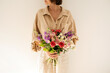 © Floral Deco - Young beautiful woman in beige muslin shirt hold bouquet of colourful roses flowers against white wall. Beautiful holiday celebration floral composition