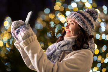 Poster - holidays, communication and people concept - portrait of beautiful happy smiling young woman taking selfie with smartphone over christmas lights in winter city