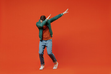 Wall Mural - Full size body length fun young brunet man 20s wears red t-shirt green jacket doing dab hip hop dance hands move gesture youth sign hide cover face isolated on plain orange background studio portrait