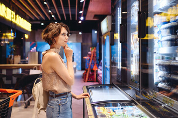 Wall Mural - Young minded woman in casual clothes shopping at supermaket greengrocery store buy choosing dairy produce cheese near refrigerator prop up chin inside hypermarket. Purchasing gastronomy food concept