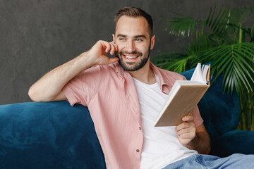 Poster - Young smiling happy satisfied dreamful man in casual clothes reading book novel look aside sitting on blue sofa at home flat indoors rest relax on weekends free time. People lounge lifestyle concept