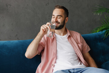 Canvas Print - Young satisfied calm man in casual clothes hold drink clear fresh pure still water from transparent glass show thumb up sit on blue sofa at home flat indoors rest on weekends. People lounge concept