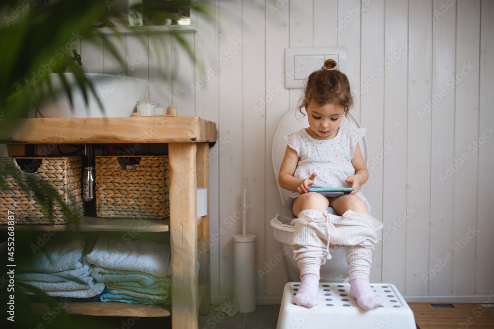Portrait of cute small sitting on toilet indoors at home, using smartphone.