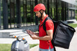 © LIGHTFIELD STUDIOS - Side view of muslim deliveryman with thermo backpack holding clipboard and pen near blurred scooter outdoors