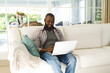 © WavebreakMediaMicro - Smiling african american man using laptop and sitting on couch in living room
