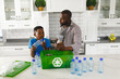 Happy african american father and son in kitchen talking and sorting plastic waste for recycling