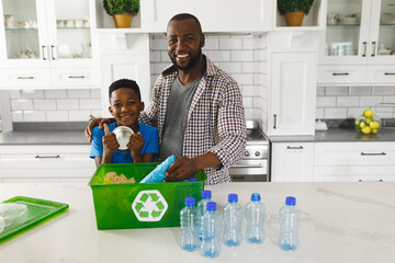 Portrait of happy african american father and son in kitchen sorting rubbish for recycling