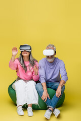 Wall Mural - interracial and middle aged couple in vr headsets sitting in bean bag chair and gaming on yellow