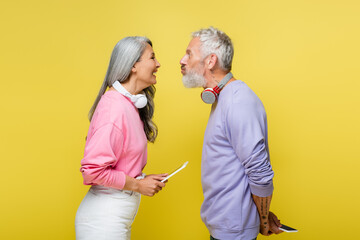 Wall Mural - side view of funny multiethnic and middle aged couple in wireless headphones looking at each other and grimacing isolated on yellow