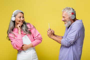 Wall Mural - happy middle aged and multiethnic man in wireless headphones taking photo of cheerful wife isolated on yellow