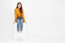 Portrait Of Young Asian Woman Sitting On White Box Isolated Over White Background, Full Body Composition