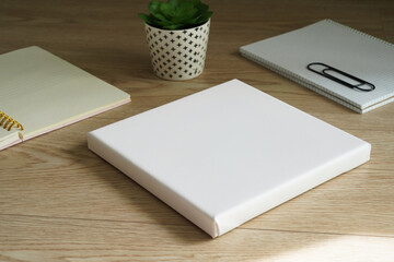 White empty square canvas for painting lies on a wooden table next to two notes and a small flower. Mockup canvas (982)