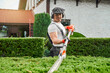 Happy male gardener in safety mask and gloves using electric hedge trimmer while working on backyard. Caucasian man cutting overgrown bushes outdoors