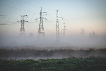  A power line passes over the river in Latvia. Morning shot in the haze on the background of the forest. Lots of pylons in meadow looks like big giants