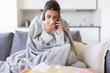 © deniskomarov - Sick female calling doctor, blowing, wiping running nose, sneezing in tissue, sitting on sofa. Ill  woman consulting with practitioner on phone, covered with blanket, having influenza flu symptoms