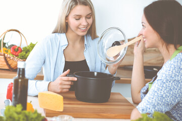 Wall Mural - Two women friends looking into the dark pot with a ready meal and taste new recipes while sitting at the kitchen table. Healthy meal cooking concept