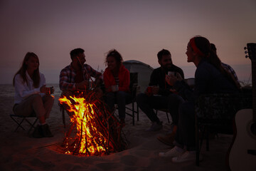 Canvas Print - Group of friends gathering around bonfire on beach in evening. Camping season