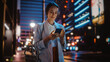 © Gorodenkoff - Beautiful Young Woman Using Smartphone Walking Through Night City Street Full of Neon Light. Smiling Thoughtfully Female Using Mobile Phone, Posting Social Media, Online Shopping, Texting.