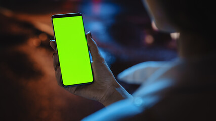 Beautiful Woman using Chroma Key Smartphone while Walking Through Night City Street Full of Neon Light. Female Using Green Screen Mobile Phone. Over the Shoulder Close-up Shot