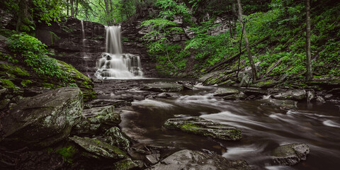 Natural Waterfall in a forest with green foliage surrounded by large rocks along a flowing river - Long exposure photography - camping hiking - forest landscapes -  trees with green leaves