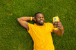 © shunevich - Happy young adult man laying on the grass, using smartphone, browsing internet. Joyful african american male relaxing, taking a break after workout outdoors, having a video call from friends, smiling