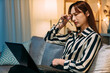 overworked asian businesswoman holding eyeglasses is rubbing her sore eyes while working overtime with a laptop on the couch in the living room at home.