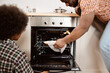 © pressmaster - Young African man taking hot baked apple pie out of electric oven in front of his little son