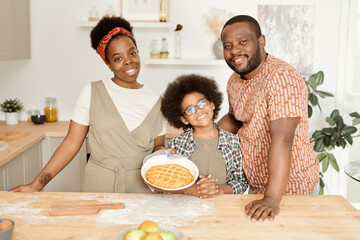 Wall Mural - Happy young African family of three with fresh baked apple pie standing by kitchen table