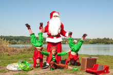 Children Elves Stand On Their Hands In The Summer Against The Backdrop Of The River Of The Sea In Costumes With Santa Claus Inflatable Cooges And Sleds In The Summer For Christmas And New Year's