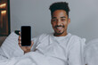 © ViDi Studio - Young happy african american man in nightwear hold mobile cell phone with blank screen workspace area lying in bed rest relax spend time in bedroom lounge home in own room house wake up in good day.