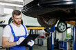 Young professional technician mechanic man wears denim overalls use hold clipboard papers document estimate stand near car lift check technical condition work in vehicle repair shop workshop indoors.