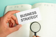 © Maks_Lab - a man's hand holds a white card with the text Business Strategy above an open paper notebook and a magnifying glass in a black frame